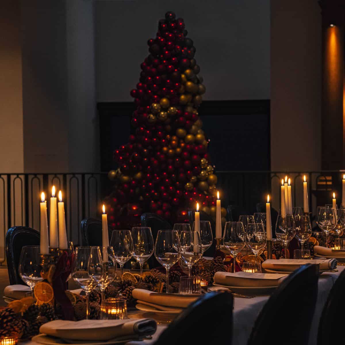 Long banquet table set for a Christmas party at Town Hall, with lit candles, dried orange and pine cone garlands, and a tree of red and gold baubles in front of the mezzanine